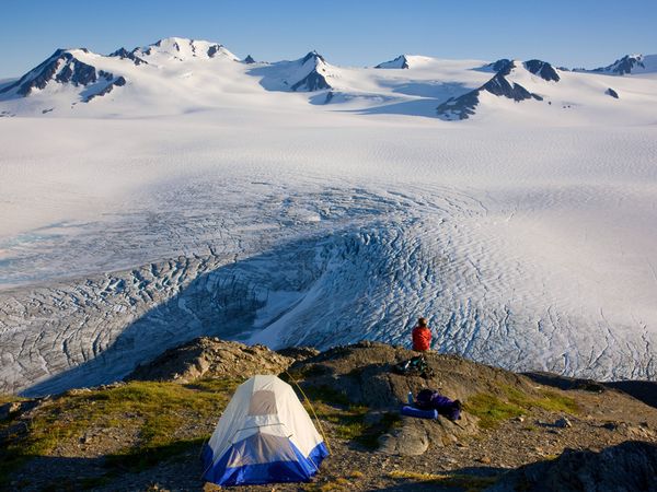snowy peaks and white ice field with a bare peak with a lone camper and tent on top