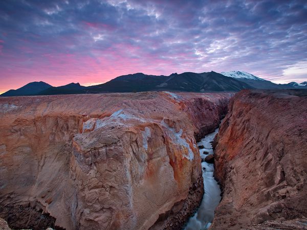 a steep, narrow river canyon cuts through reddish orange rock with mountains and a pink sunset behind