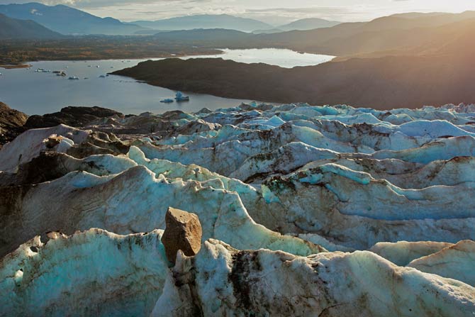 a boulder sits high atop a glacier with water at the base of the glacier and mountains in the background