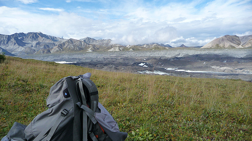 A backpack sits in a foreground of grass while the Alaska range, Mt. McKinley snow capped peaks and Denali rivers are captured in the background - Princess Lodges