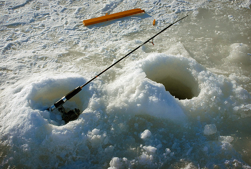 Two holes are cut in ice with a fishing pole standing upright out of the hole on the left. 