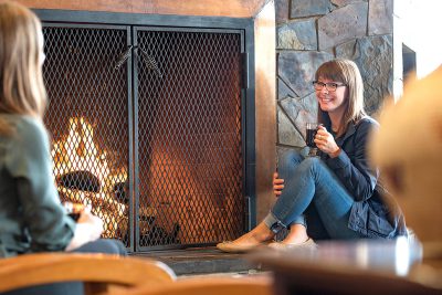 A woman sips coffee in front of a fireplace while talking to another woman.